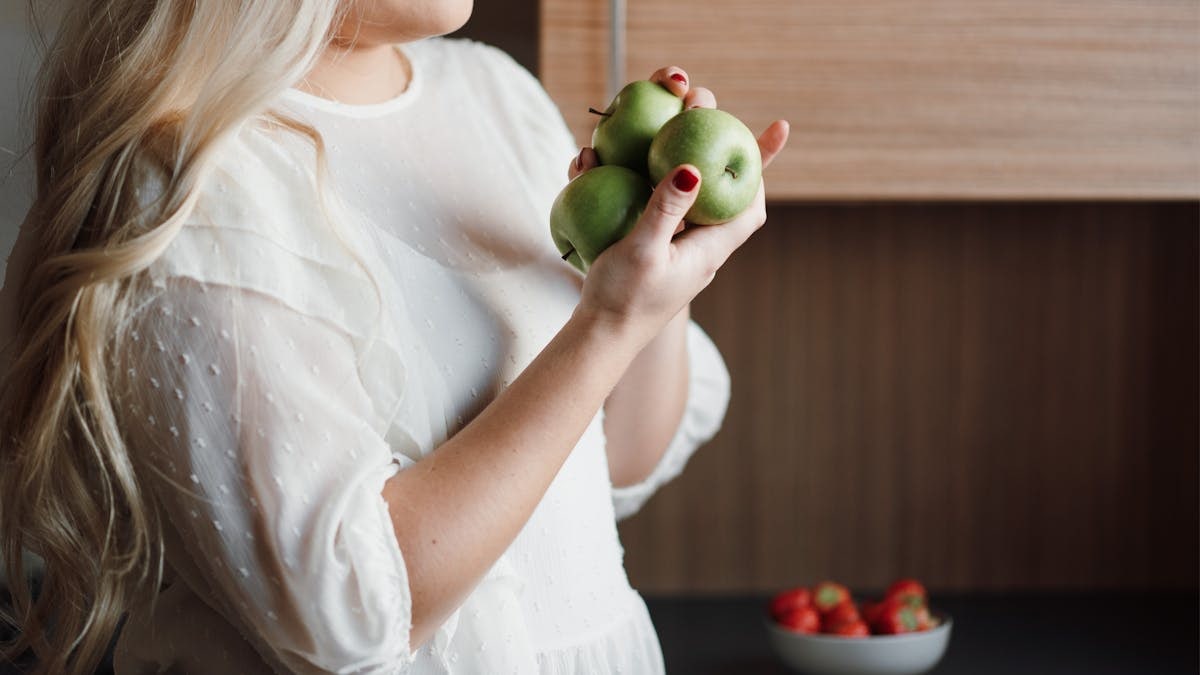Side view of crop unrecognizable positive lady with long blond hair smiling while holding fresh green apples in hands standing near table with bowl of ripe strawberries